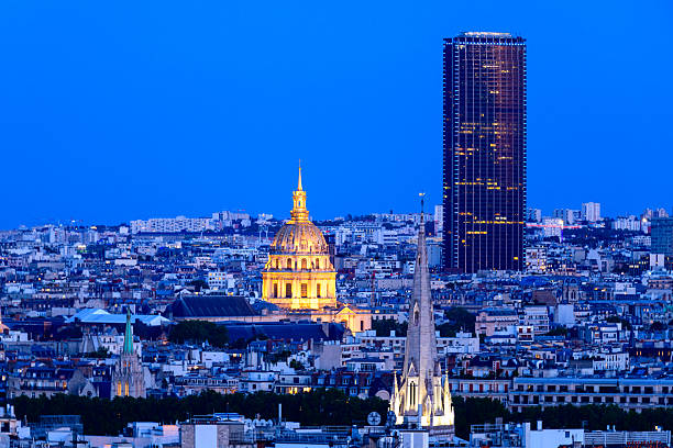 Evening cityscape of Paris featuring the glowing dome of Les Invalides, the modern silhouette of Montparnasse Tower, and the spire of the American Cathedral against a deep blue sky.