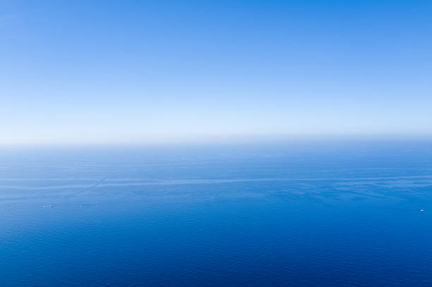 Minimalist seascape with calm, deep blue Mediterranean water stretching to a clear sky, creating a tranquil and expansive atmosphere. Subtle gradients of blue and faint boat trails evoke openness and serenity.