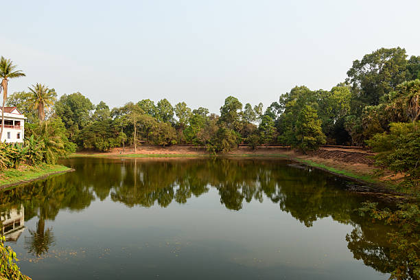Calm surface of a rectangular moat bordered by lush green trees and tropical vegetation near Bakong in Cambodia. The tranquil scene features clear reflections, earthy banks, and soft daylight, evoking a peaceful rural atmosphere.