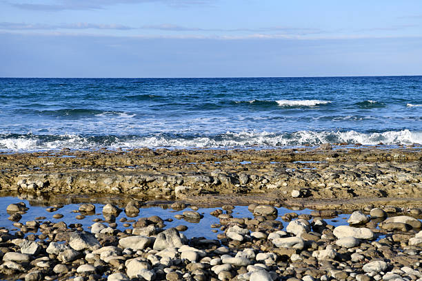 A wide horizontal shot of the Mediterranean coast featuring a layered rock formation acting as a natural barrier between the blue ocean waves and a calm, shallow tide pool filled with pebbles in the foreground.
