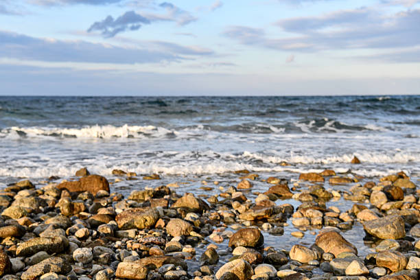 A wide shot of a rocky coastline with small waves washing over a variety of smooth stones. The horizon meets a soft blue sky with scattered clouds, creating a peaceful maritime atmosphere.