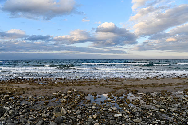 A wide coastal view featuring gentle blue waves breaking over a rocky shoreline. The horizon meets a soft, cloud-filled sky, creating a tranquil and scenic maritime atmosphere.