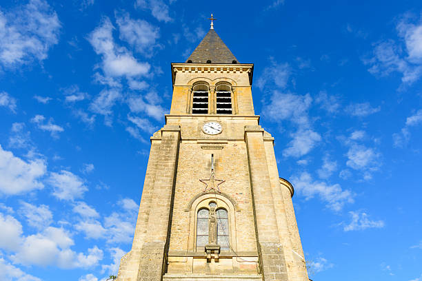 Upward view of a historic stone church bell tower featuring arched windows and a prominent clock, set against a vivid blue sky with scattered clouds in Saint-Honore-les-Bains.