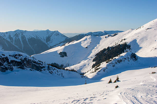 Brilliant midday sun shines in a clear blue sky above a panoramic view of the snow-covered Alpine mountain peaks. In the foreground, patches of snow and brown vegetation cover a mountain ridge. A thin airplane contrail is visible in the upper right corner of the sky. The mountain range stretches across the horizon with multiple layers of peaks fading into the distance.