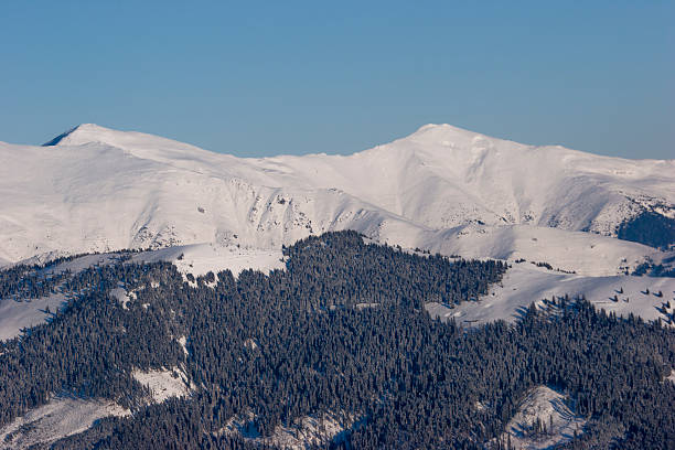 A landscape shot of a massive, snow-blanketed mountain range beneath a brilliant, deep blue sky. The foreground highlights the unique wind-blown textures and ripples in the snow, leading the viewer's eye toward several sharp, sunlit peaks. The image evokes a sense of cold, quiet majesty and the vastness of the alpine wilderness, perfect for travel, adventure, and nature-themed projects.