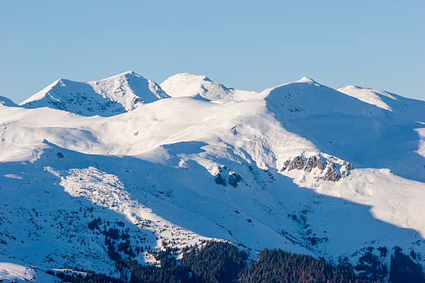 A landscape shot of a massive, snow-blanketed mountain range beneath a brilliant, deep blue sky. The foreground highlights the unique wind-blown textures and ripples in the snow, leading the viewer's eye toward several sharp, sunlit peaks. The image evokes a sense of cold, quiet majesty and the vastness of the alpine wilderness, perfect for travel, adventure, and nature-themed projects.