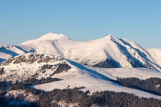 A landscape shot of a massive, snow-blanketed mountain range beneath a brilliant, deep blue sky. The foreground highlights the unique wind-blown textures and ripples in the snow, leading the viewer's eye toward several sharp, sunlit peaks. The image evokes a sense of cold, quiet majesty and the vastness of the alpine wilderness, perfect for travel, adventure, and nature-themed projects.