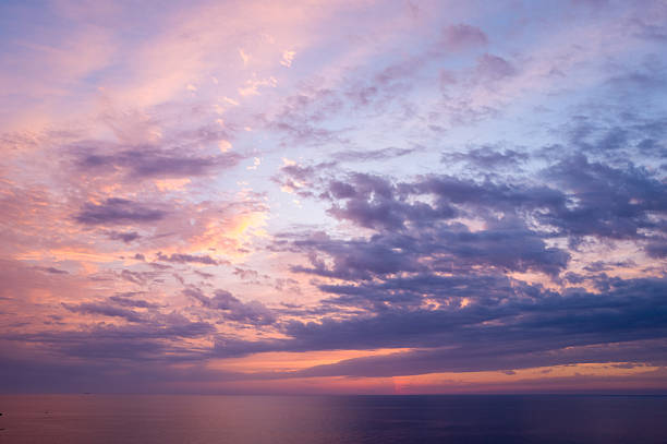 Colorful bands of lavender, pink, and blue clouds stretch across the evening sky above the calm Mediterranean waters. The tranquil scene is bathed in soft, glowing twilight, creating a serene and atmospheric mood.