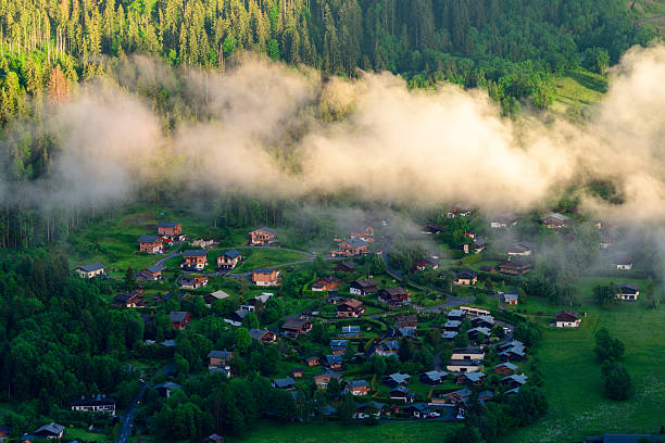 Low clouds drift above a cluster of traditional chalets surrounded by lush green forest at sunrise in Les Houches, creating a peaceful and atmospheric mountain setting.