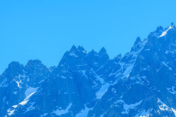 Rugged, jagged summits of Aiguille de Blaitiere rise sharply against a vivid blue sky, with patches of lingering snow accentuating the dramatic alpine rock formations. Bright daylight highlights the textured surfaces and steep ridgelines.
