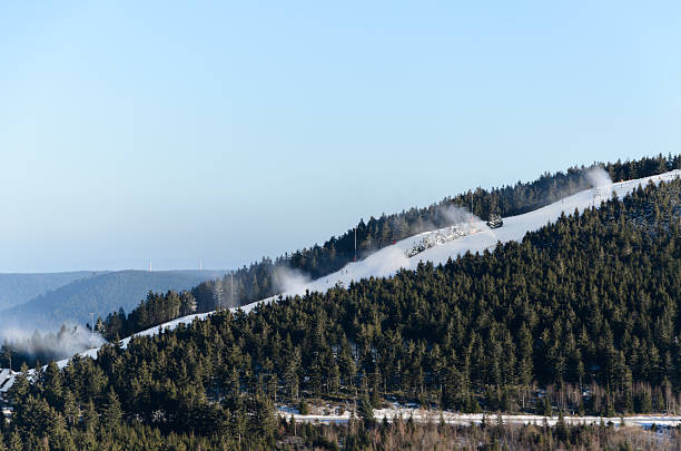 Snow cannons spray artificial snow on a white ski slope cutting through a dense green pine forest under a clear blue winter sky.