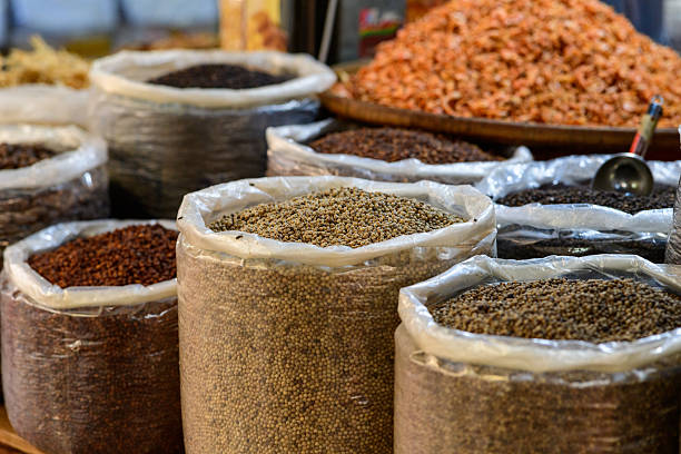 Large sacks of assorted whole spices, including white peppercorns and black pepper, are displayed alongside a mound of dried shrimp at a traditional market in Kampot, Cambodia. The image highlights natural textures, earthy colors, and the abundance of local ingredients in a Southeast Asian food market.