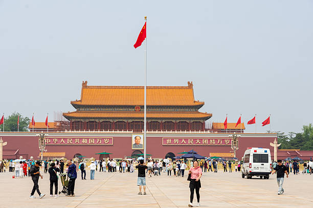 Beijing, China, October 10th, 2023: A wide view of Tiananmen Gate in Beijing, China, set against a bright blue sky. Red walls, orange roofs, and fountains line a busy street with cars and flags.