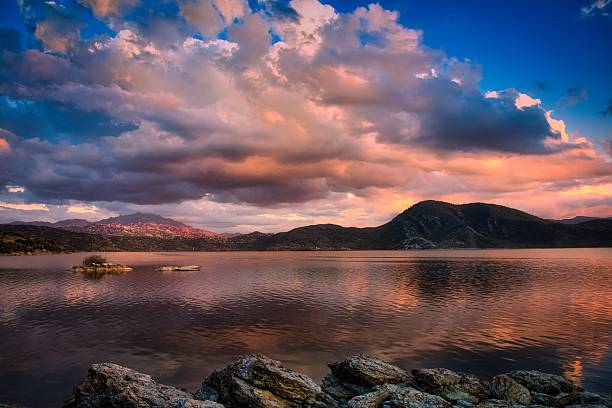 Spectacular sunset at Lake Bafa. Mighty cumulus clouds glowing pink from below are reflected in the calm water. Wide-angle view with rugged rocks in the foreground and the majestic Latmos massif on the horizon.