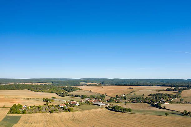 Wide aerial view of a small countryside village surrounded by expansive golden farmland, scattered trees, and distant woodland under a vivid blue sky. The open landscape and gentle hills create a sense of spaciousness and rural tranquility in central France.
