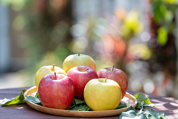 Six premium Japanese apples displayed on a rustic wooden plate outdoors, featuring fresh organic fruit with vibrant colors, natural light, and shallow depth of field for healthy food concepts.