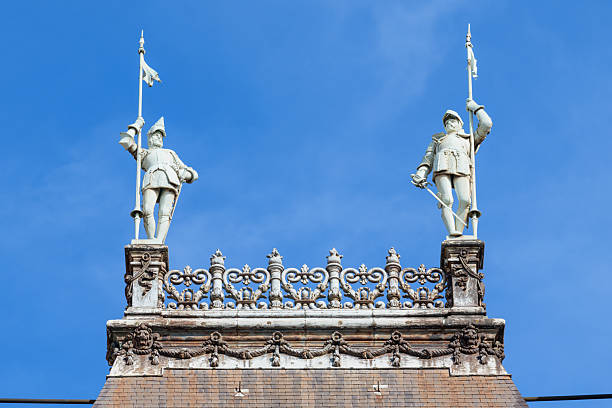 Pair of stone renaissance guard statues holding spears stand atop a decorative ironwork balustrade on the rooftop of Hotel de Ville in Paris, highlighted by bright daylight against a vivid blue sky.