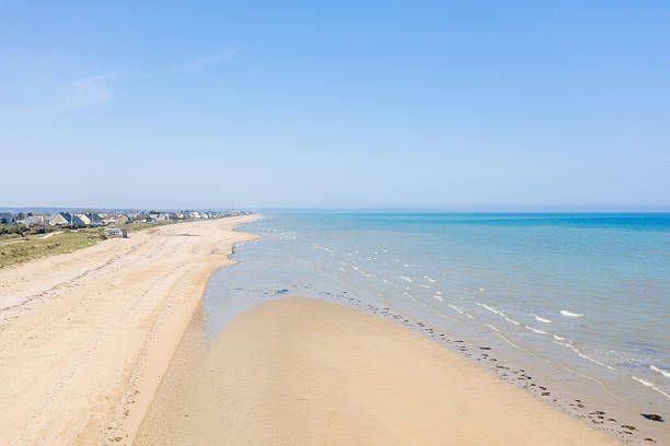 Expansive view of Utah Beach with pale sand, gentle waves, and a row of distant houses along the grassy dunes. Clear blue sky and bright sunlight create a peaceful, open coastal scene.