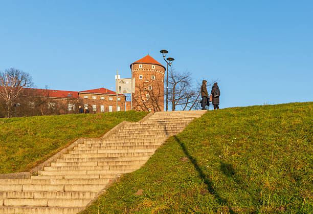 Krakow, Poland – December 13, 2025: Stone steps rise from the river embankment toward the grassy slope near Wawel Hill, with the historic Wawel defensive tower visible above during a bright winter afternoon.