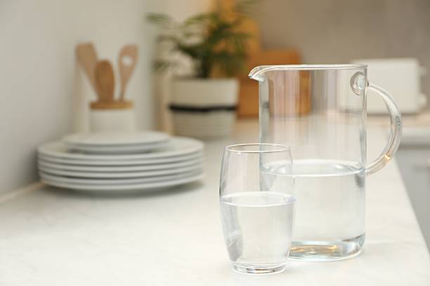 Jug and glass with clear water on white table in kitchen, space for text