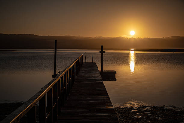 A wooden jetty glows in warm golden light as the sun sets behind the surrounding mountains of a tranquil lagoon. The calm water reflects the rich colors of the sky, creating a serene and picturesque landscape.