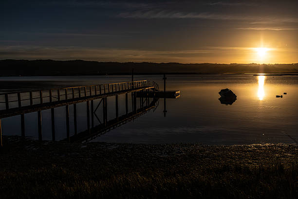A wooden jetty glows in warm golden light as the sun sets behind the surrounding mountains of a tranquil lagoon. The calm water reflects the rich colors of the sky, creating a serene and picturesque landscape.