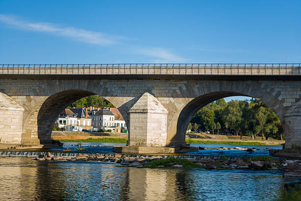 Historic stone bridge with wide arches spans the Loire river, casting shadows on the calm water below. Traditional riverside houses and lush trees frame the scene under a vivid blue sky in La Charite sur Loire.