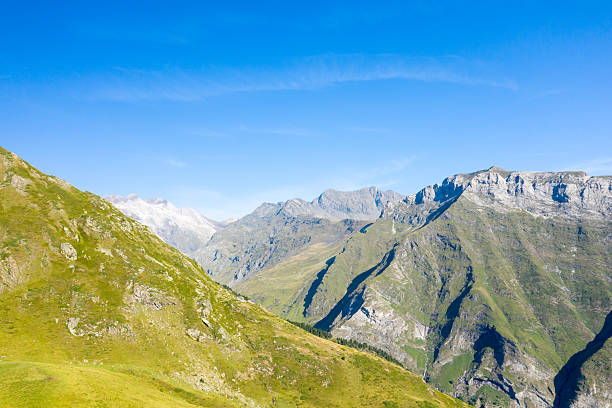 Wide view of sunlit grassy hills and rugged stone ridges under a brilliant blue sky near Gavarnie Gedre in the Pyrenees. The scene features dramatic elevation and crisp natural textures in clear summer light.