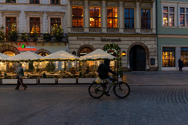 Krakow, Poland – December 3, 2025: A cyclist rides across the Main Market Square past illuminated restaurant terraces. Historic buildings and Christmas decorations frame the calm winter evening in the Old Town.