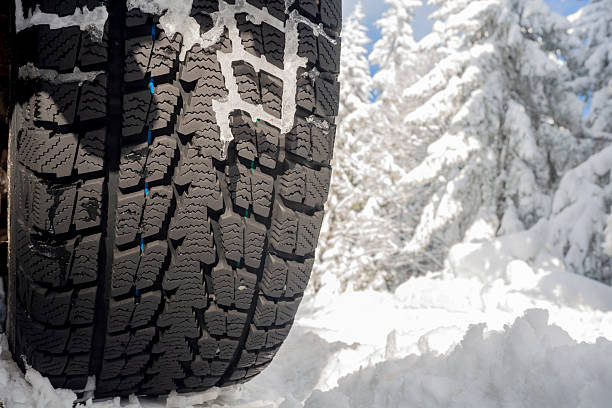 A detailed, low-angle close-up of a vehicle's winter tire tread heavily packed with snow. The image highlights the deep grooves and sipes of the rubber designed for maximum traction on icy surfaces. In the background, a blurred snowy road and misty evergreen forest emphasize a cold, high-altitude environment. This shot is ideal for automotive advertising, road safety articles, and winter travel guides.