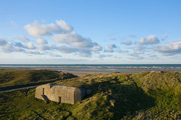 A concrete World War II bunker rests in windswept grassy dunes overlooking the wide sandy shore and calm sea at Utah Beach, under a sky dotted with soft clouds. The scene blends rugged historic architecture with natural coastal textures and open space.