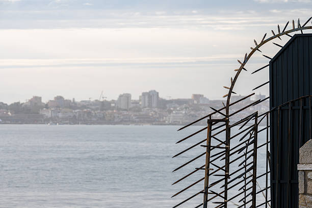 Estoril Portugal December 17 2025. A spiked metal fence lines a coastal edge with the Atlantic Ocean and Estoril shoreline visible in the background.