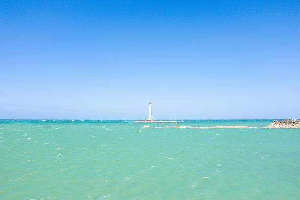 A solitary lighthouse stands on a distant rocky islet, surrounded by turquoise-green sea and under a cloudless blue sky near Cap de la Hague. The image features striking minimalism, vibrant color, and a tranquil mood.