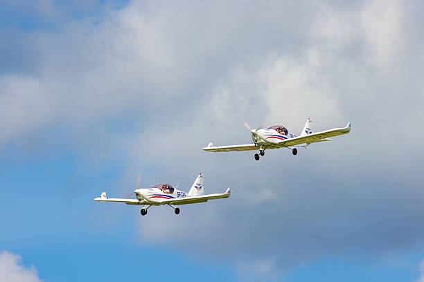Suwalki, Poland, June 26 2025 (Odlotowe Suwalki 2025) - Two white Aero AT-3 R100 light aircraft are captured in flight, flying in formation against a backdrop of a partially cloudy sky. Both planes feature distinctive red and blue stripes along their fuselages and wings, and are branded with 'AEROKLUB WARSZAWSKI' (Warsaw Aeroclub) markings. The pilots are visible in the cockpits, suggesting an active flight, possibly during an airshow, training exercise, or a recreational flight. The image provides a detailed view of these specific aircraft models in dynamic motion.