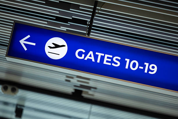 Suspended airport wayfinding sign with a blue background and white text, directing passengers toward gates 10–19 with a left-pointing arrow and airplane icon. The modern ceiling design with linear panels and integrated lighting enhances the professional atmosphere of the terminal. From a business concept perspective, the image represents clarity, organization, and efficiency in customer experience—key values in aviation, travel, and logistics industries. It can be used to illustrate themes of global mobility, infrastructure design, passenger guidance, and operational excellence. Suitable for business presentations, travel blogs, corporate reports, or stock photography collections, the image conveys trust, direction, and streamlined service in modern transportation environments.