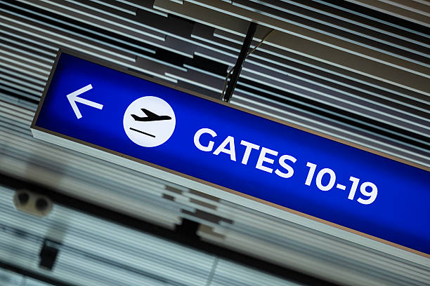 Suspended airport wayfinding sign with a blue background and white text, directing passengers toward gates 10–19 with a left-pointing arrow and airplane icon. The modern ceiling design with linear panels and integrated lighting enhances the professional atmosphere of the terminal. From a business concept perspective, the image represents clarity, organization, and efficiency in customer experience—key values in aviation, travel, and logistics industries. It can be used to illustrate themes of global mobility, infrastructure design, passenger guidance, and operational excellence. Suitable for business presentations, travel blogs, corporate reports, or stock photography collections, the image conveys trust, direction, and streamlined service in modern transportation environments.