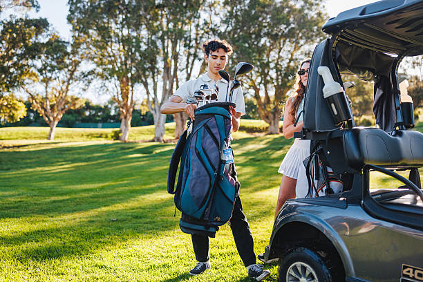 Young Caucasian couple happily preparing golf bags beside a cart on a sunlit golf course, engaging in joyful setup before their game.