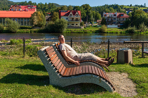 Side angle of a man resting on a curved wooden sun lounger near a riverbank. Warm daylight, green surroundings and calm water evoke slow travel, summertime отдых, mindfulness, and outdoor leisure.