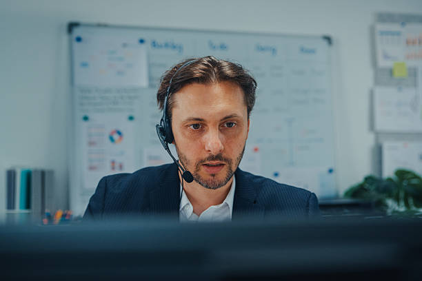 istock Close-up young male customer service agent wearing a headset talks to a client while working at computer in call center office. Sales support, contact center support and professional communication. 2254921476