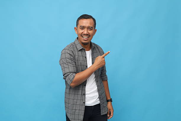 istock Happy smiling Asian man looking at camera while pointing to the side in empty space, isolated on blue background. 2254922433