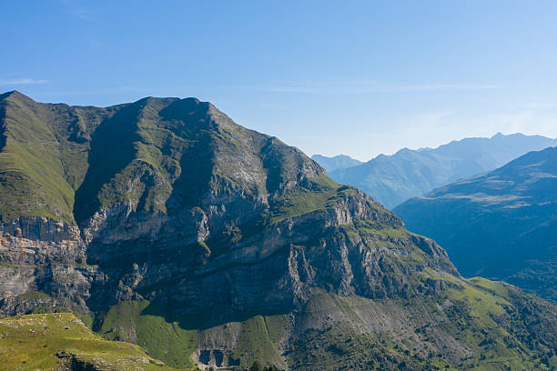 Majestic green and rocky cliffs rise sharply under a clear blue sky in the Pyrenees near Gavarnie Gedre. Layers of distant peaks fade into the horizon, creating a tranquil and expansive alpine scene.