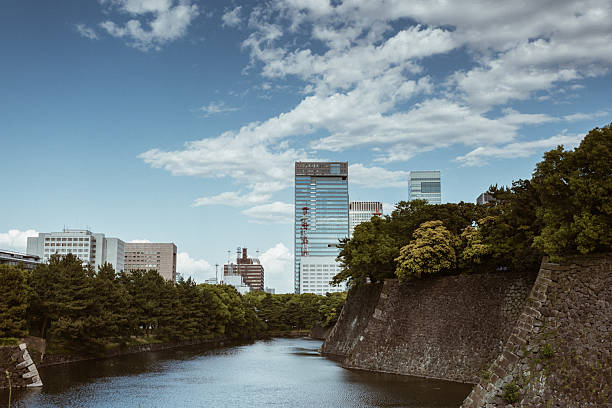 Serene view of a historic city moat with ancient stone walls and vibrant green trees framing the calm blue waters. In the background, towering modern skyscrapers rise against a partly cloudy sky, capturing the blend of historical architecture and contemporary urban skyline. Ideal for illustrating urban harmony between tradition and modernity.