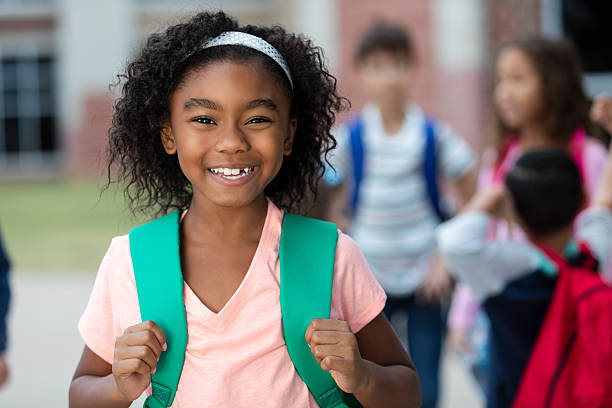 istock Joyful young Black girl with backpack smiles at camera, other students blurred in background 2254930945