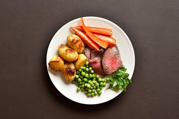 Sunday Roast with roasted beef, carrots, crispy potato, green pea and yorkshire pudding. Traditional british dish. Brown stone background, top view, flat lay