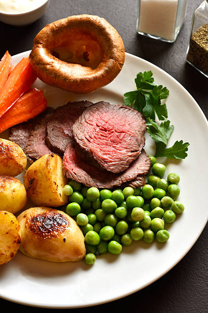 Sunday Roast with roasted beef, carrots, crispy potato, green pea and yorkshire pudding on white plate on brown stone background. Traditional british dish. Close up view