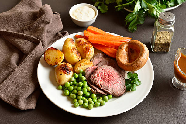 Sunday Roast with roasted beef, carrots, crispy potato, green pea and yorkshire pudding on white plate on brown stone background. Traditional british dish. Close up view