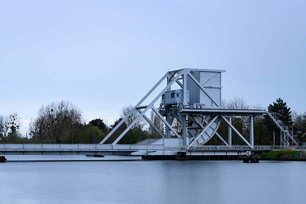 Contemporary white steel drawbridge spans a tranquil river, surrounded by bare trees and soft daylight under an overcast sky, creating a serene and industrial atmosphere.