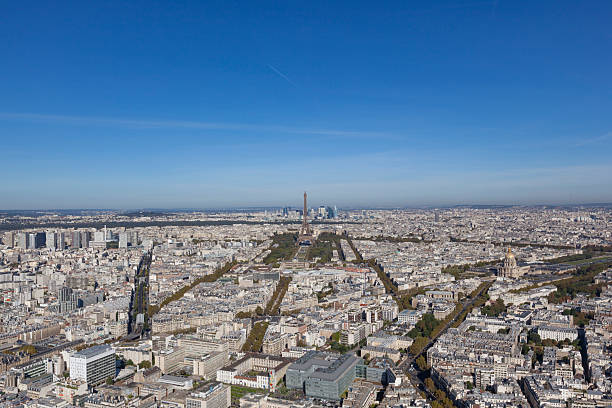 Wide panoramic view of Paris, France, showing dense urban cityscape with the Eiffel Tower as central landmark. Aerial perspective over residential buildings, tree-lined avenues, and historic architecture under a clear blue sky. Horizontal composition with extensive detail and depth.