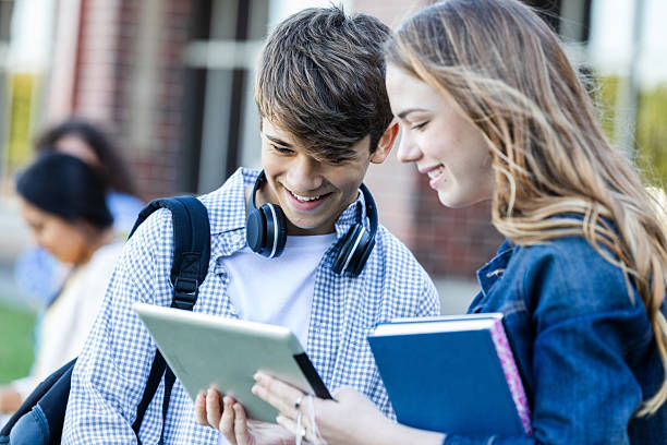 Smiling teenagers outdoors, looking at a tablet together, with a book and headphones.