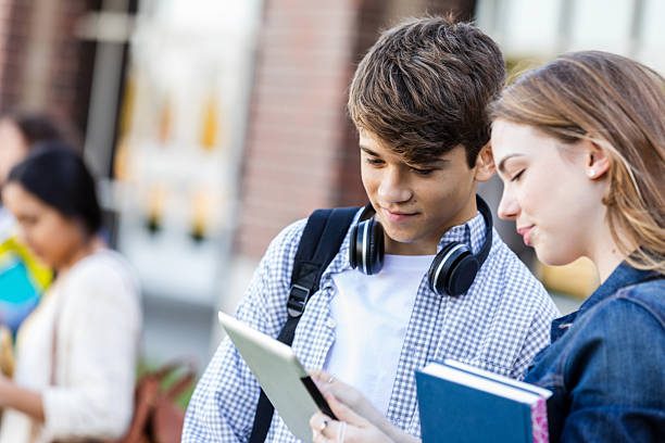 Two young students collaborate on a digital tablet and books, engrossed in their studies on campus.
